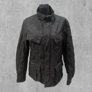 Svart quiltad jacka från Barbour - Snygg svart quiltad jacka från Barbour med hög krage och knappar. Jackan har långa ärmar och praktiska fickor framtill. Perfekt för kyligare dagar.