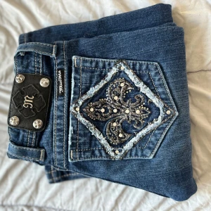 Blå jeans från Missme - Snygga blå jeans från Miss Me med unika broderade detaljer och pärlor på bakfickorna. Jeansen har en klassisk femficksdesign och en lågmidjad passform. Perfekta för dig som vill ha något extra i garderoben!