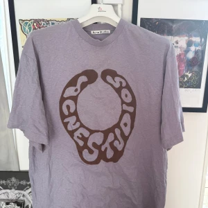 Lila t-shirt från Acne Studios - Säljer en cool lila t-shirt från Acne Studios med ett stort brunt tryck på framsidan. T-shirten har en avslappnad passform. Storlek M