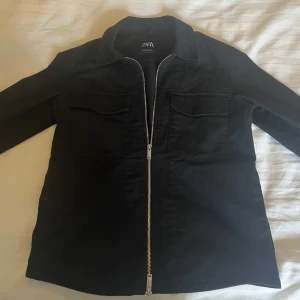 Svart jacka från Zara - Säljer en stilren svart jacka från Zara med dragkedja framtill. Jackan har långa ärmar och två bröstfickor med lock. Perfekt för en minimalistisk look.