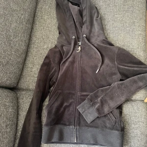 Svart juicy kofta med dragkedja - Säljer en svart juciy kofta med dragkedja och huva. Den har långa ärmar och snörning vid huvan för justering. Själva dragkedjan där det står ett "J" är lite rostig 