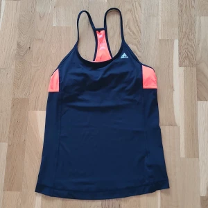 Svart och orange linnetopp från Adidas - Säljer en snygg svart och orange linnetopp från Adidas. Toppen har smala axelband och är perfekt för träning. Den är tillverkad i ett lätt och bekvämt material med Climacool-teknologi för att hålla dig sval. Adidas-loggan är tryckt på framsidan. 