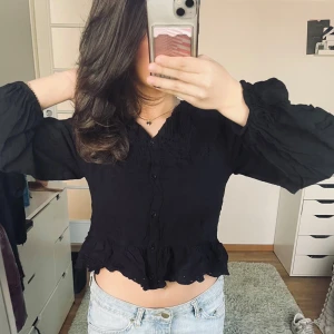 Svart blus Zara  - Säljer en elegant svart blus med volangdetaljer och knappar framtill. Blusen har långa ärmar med en lätt puffig design och en croppad passform som ger en trendig look. Perfekt för att styla med jeans eller kjol.  💕🙌🏽 från Zara 
