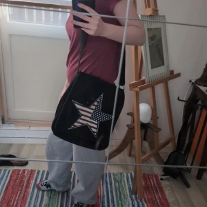 Svart axelväska med stjärndetalj från Gianni - Säljer en svart axelväska från Gianni med en stor stjärna framtill i USA-flagga-mönster. Väskan har justerbar axelrem, innerfack och stängs med knapp. Perfekt för att bära dina vardagsprylar men även skolmaterial och teknik medan samtidigt ge din outfit en cool touch.