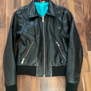 Svart skinnjacka med dragkedjor - SITTER SOM EN M. Säljer en svart skinnjacka med silverfärgade dragkedjor och ribbade muddar. Jackan har två snedställda fickor framtill och en klassisk krage. Insidan är fodrad med turkost tyg. Perfekt för dig som vill ha en stilren och tidlös look.