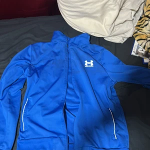 Blå träningsjacka från Under Armour - Säljer en blå träningsjacka från Under Armour med dragkedja och vita detaljer. Jackan har lång ärm, två fickor med dragkedja och logga på bröstet samt i nacken. Perfekt för träning eller sportiga dagar.