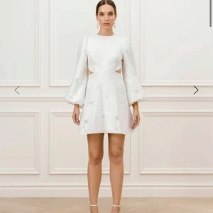 Vit kort klänning med puffärmar - Superfin vit kort klänning med puffiga långärmar och snygga cut-outs i midjan. Klänningen har rund halsringning, öppen rygg och knappar bak. Tyg med subtilt blommönster ger en lyxig känsla. Perfekt för dig som vill sticka ut med en trendig look. Ny pris 5000