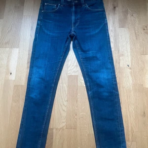 Blå skinny jeans från Nudie Jeans - Säljer ett par klassiska blå skinny jeans från Nudie Jeans. Jeansen har fem fickor, orangea sömmar och är tillverkade i 99% bomull. Snygg passform med smala ben och normal midja. Perfekta till sneakers eller boots.