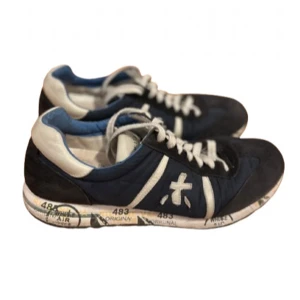 Blå och vita sneakers från Premiata - Säljer ett par blå sneakers från Premiata med vita detaljer och snörning. Skorna har en vit och mönstrad sula samt en rund tå. Materialet är en mix av mocka och syntet. Perfekta för dig som gillar stilrena och sportiga skor.