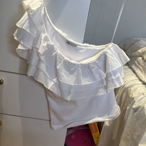 Vit volangtopp från Zara - Säljer en vit topp från Zara med bred volang runt hela ringningen. Toppen är axelbandslös och har en feminin och somrig look. Perfekt att styla med jeans eller kjol för en trendig outfit. Priset går att diskutera❤️