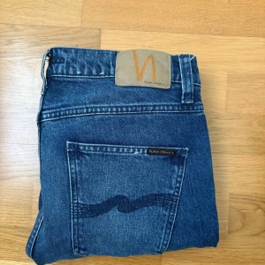 Blå jeans från Nudie Jeans - Säljer ett par klassiska blå jeans från Nudie Jeans med normal passform. Jeansen har fem fickor, synliga sömmar och Nudie-loggan på bakfickan samt läderpatch i midjan. Perfekta till vardags och har en snygg tvättad look.