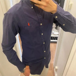 Mörkblå skjorta från Ralph Lauren - Säljer en klassisk mörkblå skjorta från Ralph Lauren med knappar hela vägen och den ikoniska röda loggan broderad på bröstet. Skjortan har lång ärm och button-down krage. Perfekt för dig som gillar stilrena plagg med snygga detaljer.