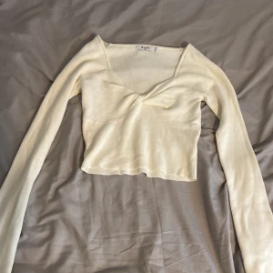 Beige/vit ribbad croppad topp från NA-KD - Säljer en  croppad topp från NA-KD med lång ärm och v-ringad front. Toppen är ribbad och har en snygg twistad detalj framtill. Perfekt för dig som gillar stilrena och trendiga plagg!