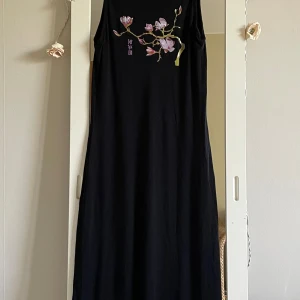 Svart långklänning med blommigt tryck - Svart ärmlös långklänning med ett fint blommigt tryck i lila och grönt framtill vid bröstet. Klänningen har en rak och enkel passform och är perfekt för en stilren look. Materialet känns mjukt och följsamt.