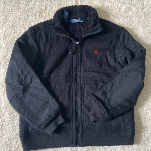 Svart fleecejacka från Polo Ralph Lauren - Svart fleecejacka med dragkedja och hög krage från Polo Ralph Lauren. Jackan har mjukt teddyfoder, förstärkta partier på ärmarna och en röd broderad logga på bröstet. Perfekt för kyliga dagar.