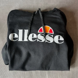 Svart hoodie från Ellesse - Säljer en svart hoodie från Ellesse med stor logga framtill i vitt, orange och rött. Tröjan har huva med dragsko och två fickor med dragkedja. Perfekt för en avslappnad stil.