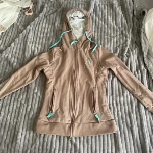 Beige hoodie från Jacson  - Säljer en beige hoodie från Equestrian Stockholm med turkosa detaljer och dragkedja. Jackan har huva med mönstrat foder, broderad logga på bröstet och text på ärmen. Perfekt för dig som gillar stilren ridstil.