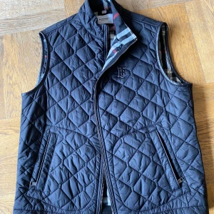 Burberry Svart quiltad väst  - Säljer en svart quiltad väst från Burberry med klassiskt rutigt foder och dragkedja framtill. Västen har två fickor med dragkedja och ett diskret broderat Burberry-logo på bröstet. Perfekt för lager-på-lager-stil. Står XL men passar mig perfekt som har storlek L