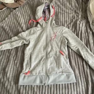 Ljusgrå hoodie från Jackson  - Ljusgrå hoodie med dragkedja, rosa snören och blommigt foder i huvan. Broderad bokstav 'J' på bröstet och texten 'Equestrian Stockholm' på ärmen. Två fickor med dragkedja och detaljer i rosa.