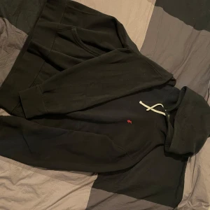 Svart hoodie från Polo Ralph Lauren - Svart hoodie från Polo Ralph Lauren med klassisk röd logga broderad på bröstet. Tröjan har huva med vita snören och en stor magficka framtill. Tillverkad i mjuk bomull, perfekt för chill dagar. Långärmad och med relaxed passform.