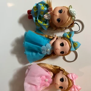 Söta docknyckelringar med klänning - Tre gulliga nyckelringar med dockor, alla med blont hår i fläta och färgglada klänningar. En har blå klänning med blommor och rödskor, en har blå klänning med rosett och en har rosa klänning med rosett. Perfekt som accessoar till väskan eller nycklarna. Du får alla tre för endast 60 kr. Helt nya.