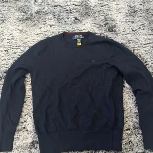 Mörkblå tröja från Polo Ralph Lauren - Klassisk mörkblå tröja från Polo Ralph Lauren med rund halsringning och diskret broderad logga på bröstet. Tröjan har långa ärmar och är tillverkad i mjuk bomull, perfekt för en stilren och enkel look.