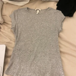 Grå tajt ribbad topp från H&M - Superenkel och snygg grå ribbad topp från H&M. Modellen är kortärmad med rund halsringning och figurnära passform. Perfekt till jeans eller shorts för en chill look. Mjuk och stretchig, passar dig som gillar basic men ändå vill ha lite struktur.