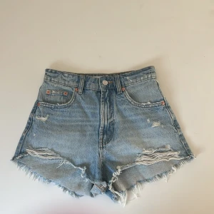 Högmidjade jeansshorts från Zara - Snygga ljusblå jeansshorts från Zara med hög midja och råa, fransiga kanter. Shortsen har slitna detaljer framtill och klassisk femficksdesign. 
