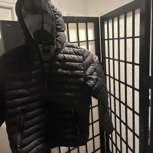 Svart dunjacka från Colmar med huva - Snygg svart dunjacka från Colmar med quiltad design och huva. Jackan har dragkedja framtill och två dragkedjefickor. Insidan är ljusgrå och axeln pryds av Colmar-logga. Perfekt för kalla och varma dagar när du vill hålla stilen.