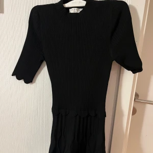Svart ribbad klänning från Sandro - Supersnygg svart ribbad klänning från Sandro Paris med kort ärm och vågig kant i midjan. Klänningen har en figurnära passform upptill och lätt utsvängd kjol med struktur. Perfekt för dig som gillar stilrena och trendiga plagg.