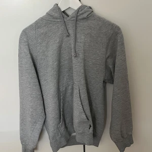 Grå hoodie med huva och ficka, M - Enkel och stilren grå hoodie i storlek M med huva och dragsnören. Tröjan har en stor magficka och långa ärmar med muddar. Perfekt för chill dagar eller när du vill ha en basic look. Materialet känns mjukt och skönt mot huden.
