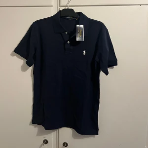 Mörkblå Polo Ralph Lauren t-shirt - Snygg mörkblå t-shirt från Polo Ralph Lauren i bomull med klassisk pikékrage och korta ärmar. Slim fit-modell med broderad vit logga på bröstet och knappar vid halsen. Perfekt för en stilren och sportig look. Pris kan diskuteras så kom gärna med prisförslag 