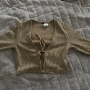 Beige ribbad bolerotopp med knyt - Säljer en beige ribbad bolerotopp med croppad passform och knyt framtill. Toppen har lång ärm och är tillverkad i mjuk bomullsmix. Perfekt att slänga över ett linne eller klänning för en chill och trendig look.