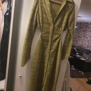 Olivgrön långklänning med struktur och knappar - Säljer en olivgrön långklänning med snygg struktur och dekorativa guldknappar framtill. Klänningen har lång ärm och en figurnära passform. Perfekt för dig som vill sticka ut med en unik look.