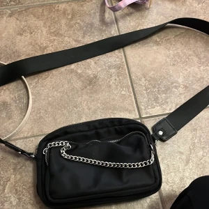 Svart axelväska från Pull&Bear - Snygg svart axelväska från Pull&Bear med bred axelrem, silverfärgad kedjedetalj och dragkedjor. Väskan har flera fack och en liten avtagbar miniväska framtill. Perfekt för att bära med sig det viktigaste och ge outfiten en cool touch.