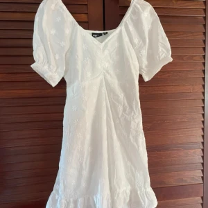 Vit blommig kortklänning från Pieces - Superfin vit kortklänning från Pieces med broderade blommor och puffärmar. Klänningen har v-ringning, dragkedja i sidan och volang nertill. Perfekt för sommarens alla tillfällen!