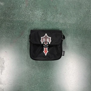 Svart axelväska med mesh och spänne - Snygg svart axelväska från Herschel med meshdetalj på framsidan och ett unikt spänne i rött och vitt. Väskan har justerbar rem och praktisk storlek för vardagens essentials. Perfekt för dig som vill ha en stilren och funktionell väska.