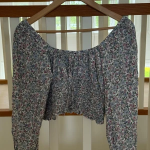 Blommig croppad blus från H&M  - Supersöt blommig blus från H&M Divided med croppad passform och långa ärmar. Toppen har smockad midja, volangkant och offshoulder-ringning. Mönstret är i pastelliga toner av blått, rosa och grönt på vit botten. Perfekt för vår och sommar!