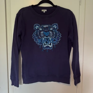 Mörkblå Kenzo sweatshirt med tiger - Kenzo sweatshirt i mörkblått med ikoniskt broderat tigerhuvud i blå och turkosa toner på bröstet. Rund halsringning, långa ärmar och ribbade muddar. Perfekt statement-plagg för dig som gillar stil och mode. Tröjan passar dej som har XS/S. Den är lite urtvättad därav priset ( ny pris 2300 ) hör av dej vid frågor och funderingar