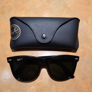 Svarta Wayfarer solglasögon från Ray-Ban - Klassiska svarta Wayfarer solglasögon från Ray-Ban med bred båge och mörka glas. Säljes med originalfodral och putsduk. Perfekt accessoar för dig som gillar tidlös och ikonisk stil.