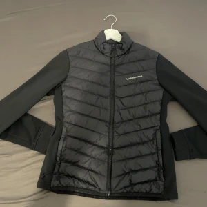 Svart hybridjacka från Peak Performance - Svart jacka från Peak Performance med quiltad front och rygg samt släta ärmar. Jackan har dragkedja framtill och broderad logga på bröstet. Perfekt för lager-på-lager och vardagsbruk.