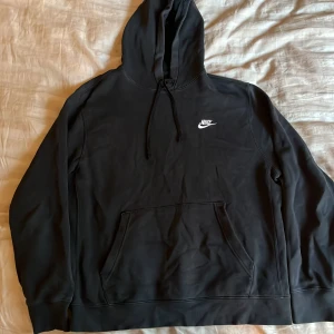Svart hoodie från Nike - Säljer en svart hoodie från Nike med vit broderad logga på bröstet. Tröjan har huva med dragsko och en stor magficka framtill. Perfekt för en avslappnad stil.