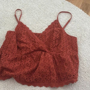 Rostfärgad croppad linnetopp med broderade detaljer - Säljer en rostfärgad croppad linnetopp med smala axelband och v-ringning. Toppen har broderade hålmönster och volangkant nedtill. Perfekt för sommaren och ger en bohemisk känsla.