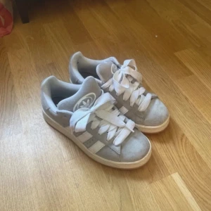 Grå Adidas campus sneakers, strl 38 - Snygga grå Adidas campus sneakers  med vita detaljer och klassiska tre ränder på sidan. Skorna har breda vita snören och rund tå. Perfekta för dig som gillar en clean och stilren look. (boxen och de gråa sko snörena finns också)
