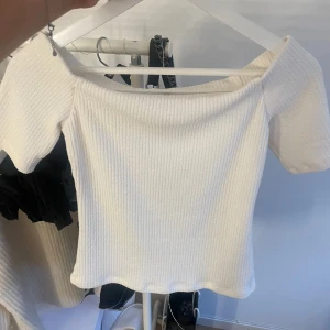 Vit ribbad offshoulder topp - Superfin vit ribbad topp med offshoulder och korta ärmar. Den har en snygg, figurnära passform och är perfekt för att lyfta vilken outfit som helst. Materialet är mjukt och stretchigt, vilket gör den både bekväm och trendig.
