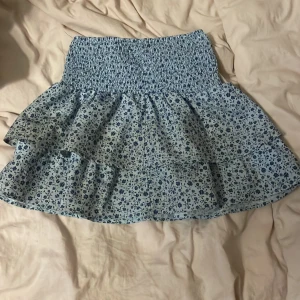 Blommig blå volangkjol - Supersöt kort kjol i ljusblått med mörkblått blommönster och bred smock i midjan. Kjolen har dubbla volanger som ger en flowy känsla och passar perfekt till varma dagar. Materialet känns lätt och luftigt.