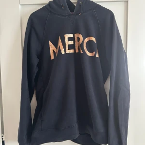 Svart hoodie med guldtext MERCI - Svart hoodie med snyggt tryck i guldtexten 'MERCI' framtill. Tröjan har huva med snörning och långa ärmar. Perfekt för en chill och avslappnad stil.