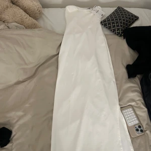 Vit axelbandslös långklänning - Säljer en elegant vit axelbandslös långklänning i glansigt material. Klänningen har en rak och stilren siluett som ger en lyxig känsla. Perfekt för dig som vill sticka ut med en minimalistisk look. Den är tight och helt oanvänd