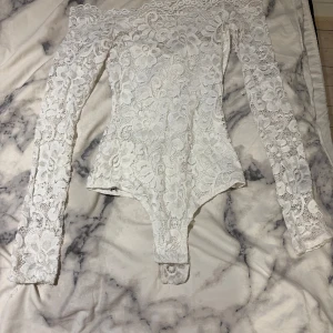Vit spetsbody från NLY One - Säljer en vit spetsbody från NLY One i storlek XS. Bodyn har långa ärmar och är helt täckt av vacker blommig spets. Perfekt för dig som vill ha en feminin och elegant topp med offshoulder-design.