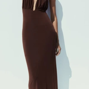 Brun långklänning med djup v-ringning - Säljer en elegant brun långklänning med djup v-ringning framtill och öppen rygg. Klänningen har en slits bak och är figurnära med ett stilrent fall. Super snygg! Endast använd 1 gång, i ny skick!